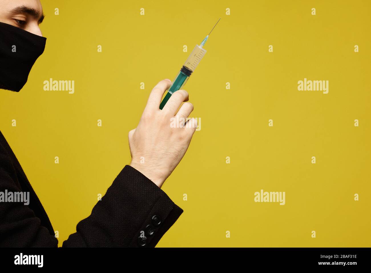 Vista laterale del bell'uomo in maschera protettiva nera che tiene una siringa in mano, isolato sullo sfondo giallo con spazio di copia per il testo. Foto Stock