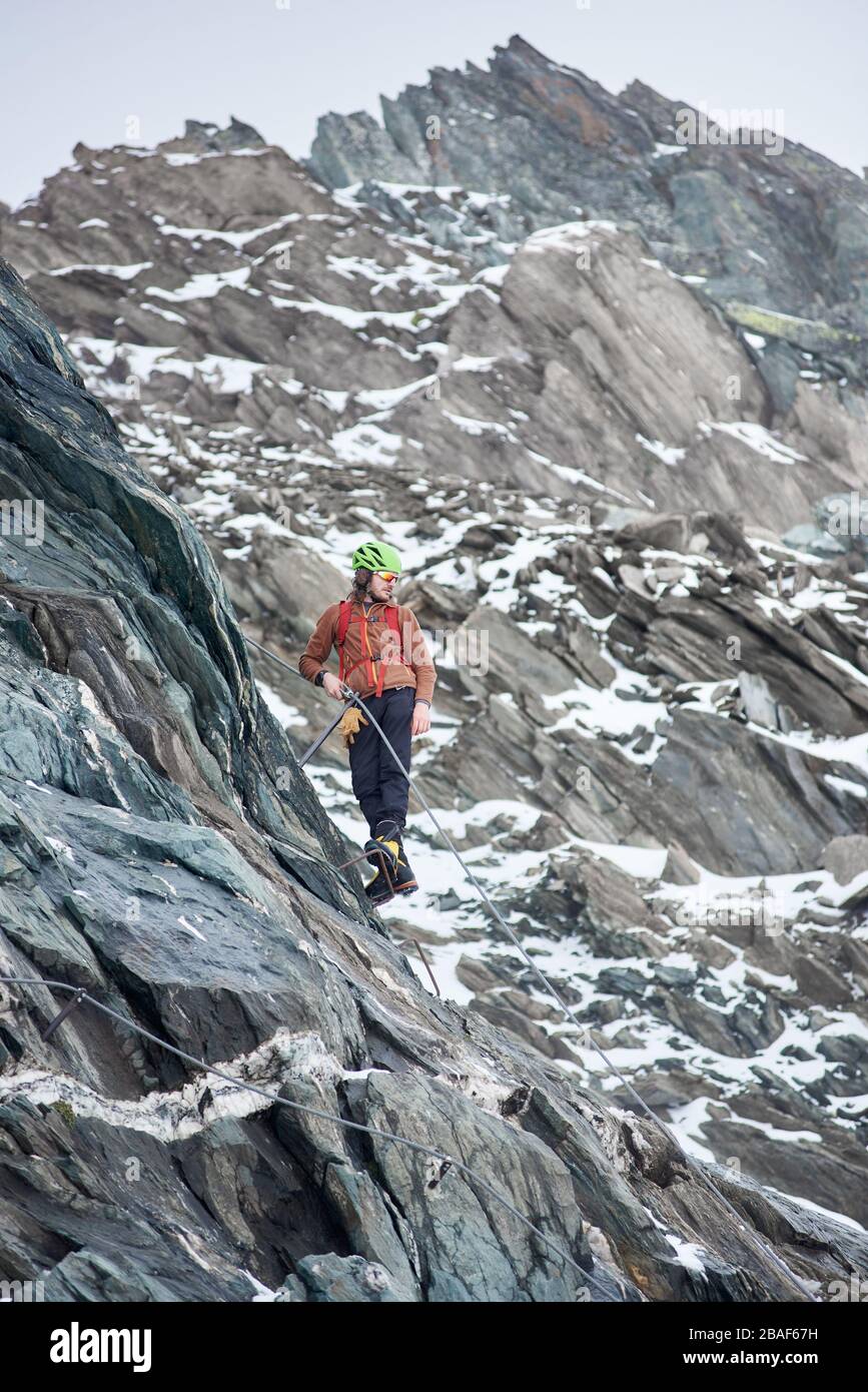 Intera lunghezza di alpinista maschile in occhiali da sole e casco di sicurezza ascendente alta montagna. Uomo arrampicatore che tiene la corda fissa e guarda verso il basso. Concetto di alpinismo, alpinismo e arrampicata. Foto Stock