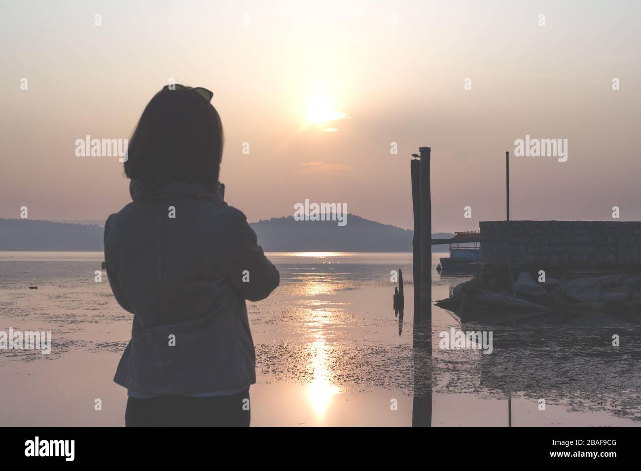 Vista posteriore della donna che guarda il molo del lago e le montagne sopra l'orizzonte al tramonto Foto Stock