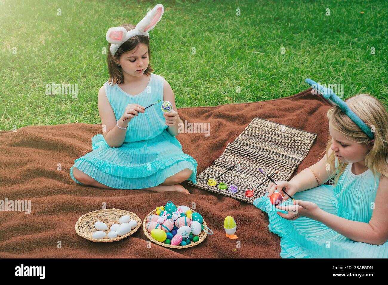Le ragazze piccole cute che portano le orecchie del coniglietto il giorno di Pasqua che si siede sulla coperta sull'erba nel giardino e che dipingano sulle uova. Foto Stock