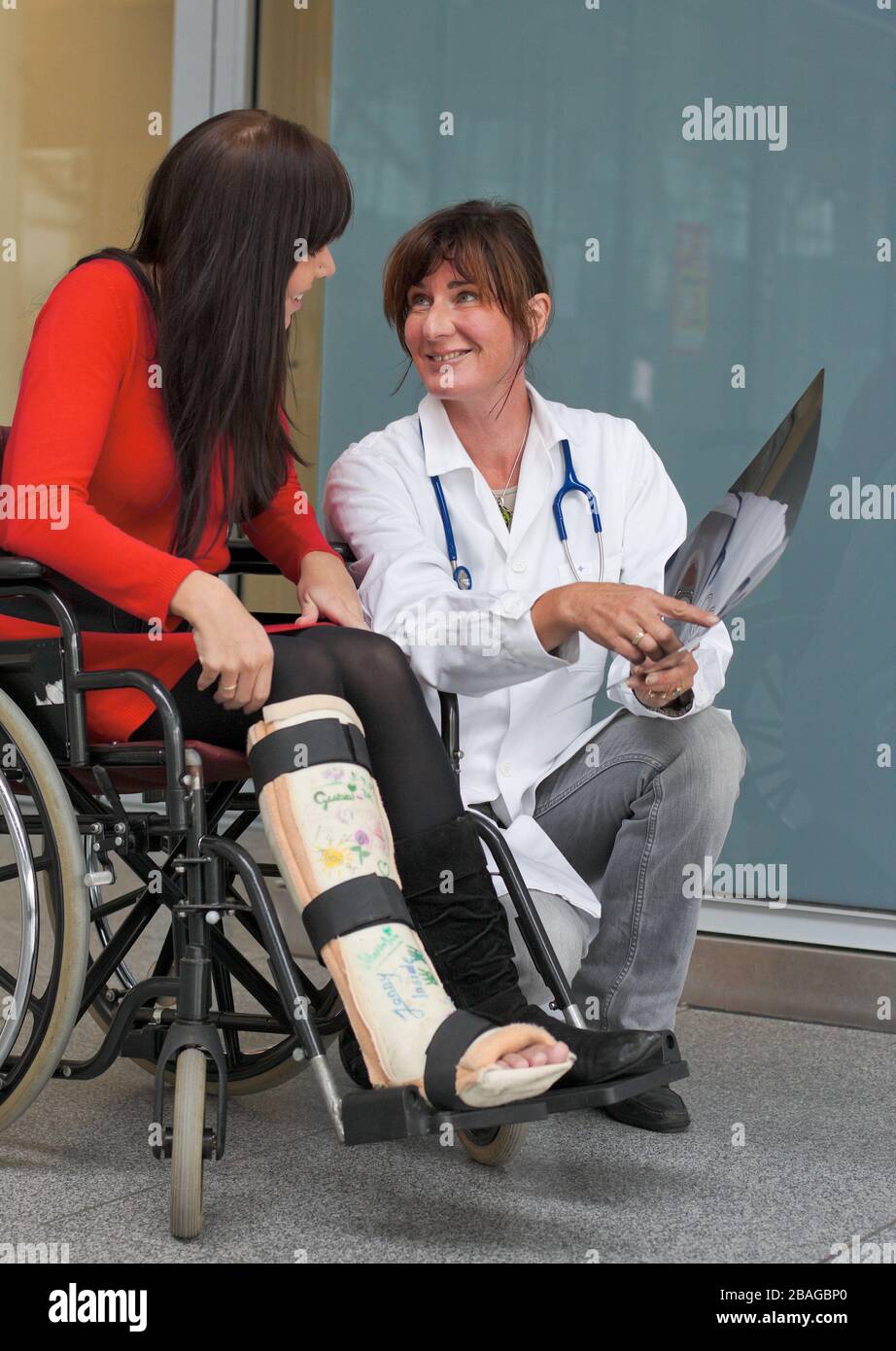 Junge Frau mit Gipsbein sitzt im Rollstuhl, MR: Si, Aerztin versorgt. sie, Foto Stock