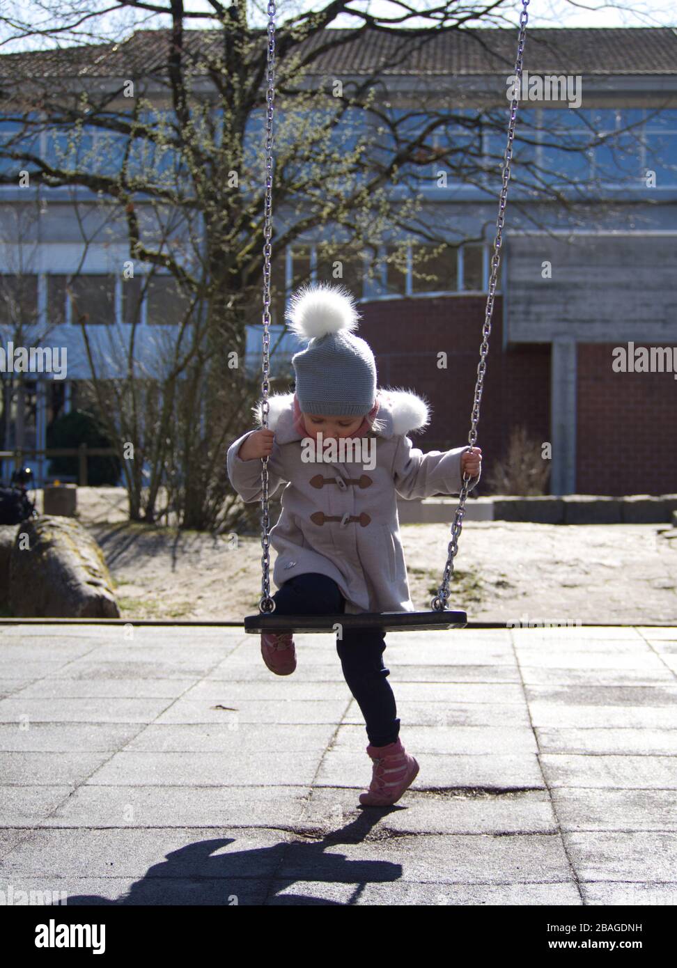 Cute piccola bionda caucasica ragazza in vestiti invernali utilizzando un'oscillazione a un bambino giocare a terra in luce solare brillante Foto Stock