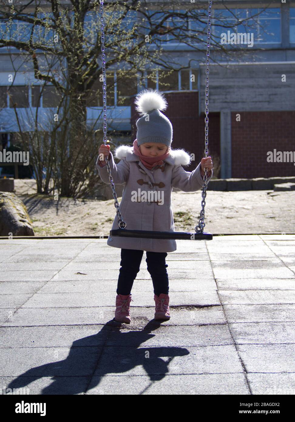 Cute piccola bionda caucasica ragazza in vestiti invernali utilizzando un'oscillazione a un bambino giocare a terra in luce solare brillante Foto Stock