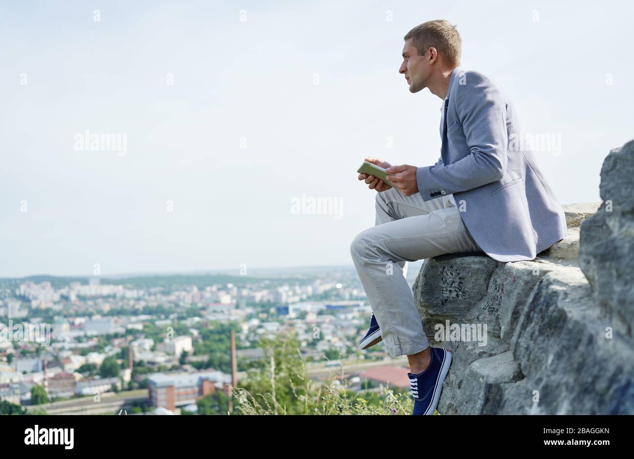 uomo d'affari con un tablet in mano siede su una roccia sullo sfondo della città e guarda in lontananza Foto Stock