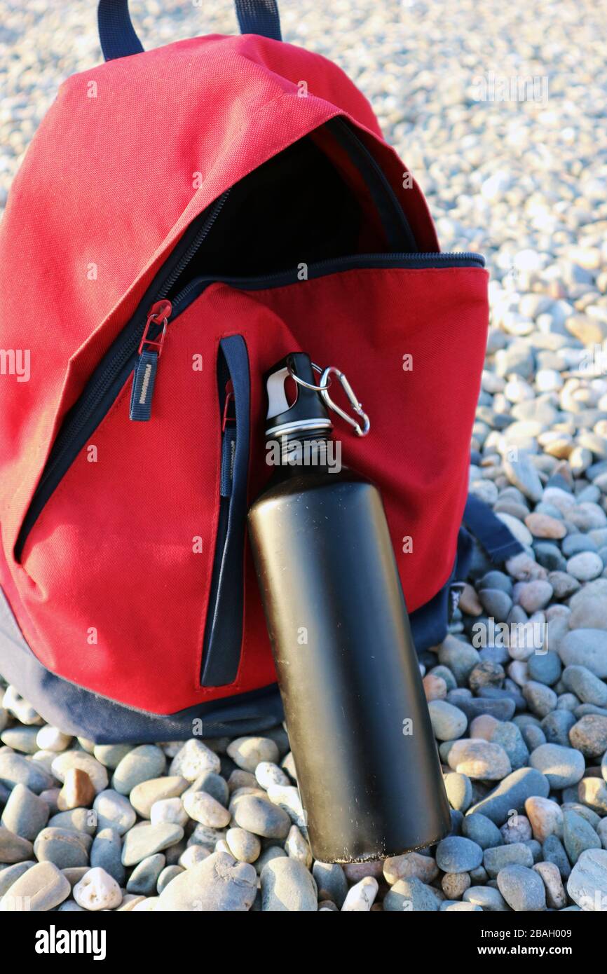 Bottiglia d'acqua in acciaio nero davanti allo zaino da viaggio rosso. Concetto di pacchetto Eco. Foto Stock