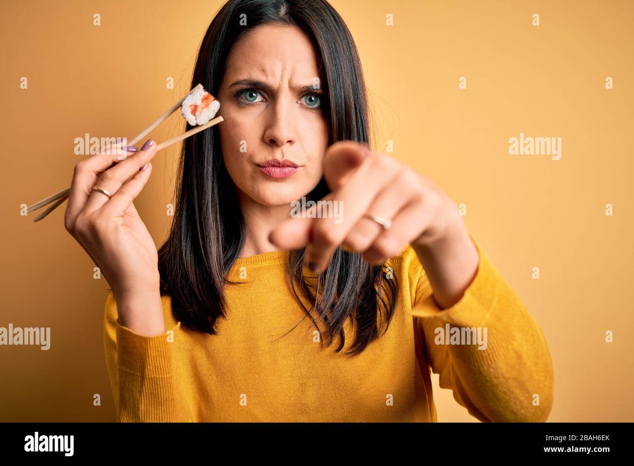 Giovane donna brunetta con occhi blu mangiare salmone maki sushi utilizzando bacchette puntate con il dito alla macchina fotografica e a voi, segno di mano, positivo e c Foto Stock