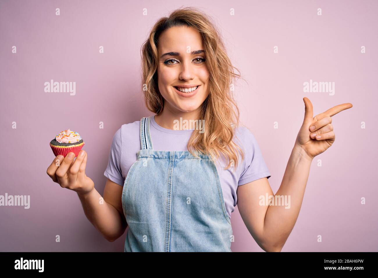 Giovane bella donna bionda eatimg cupcake cioccolato su isolato sfondo rosa molto felice indicando con mano e dito a lato Foto Stock