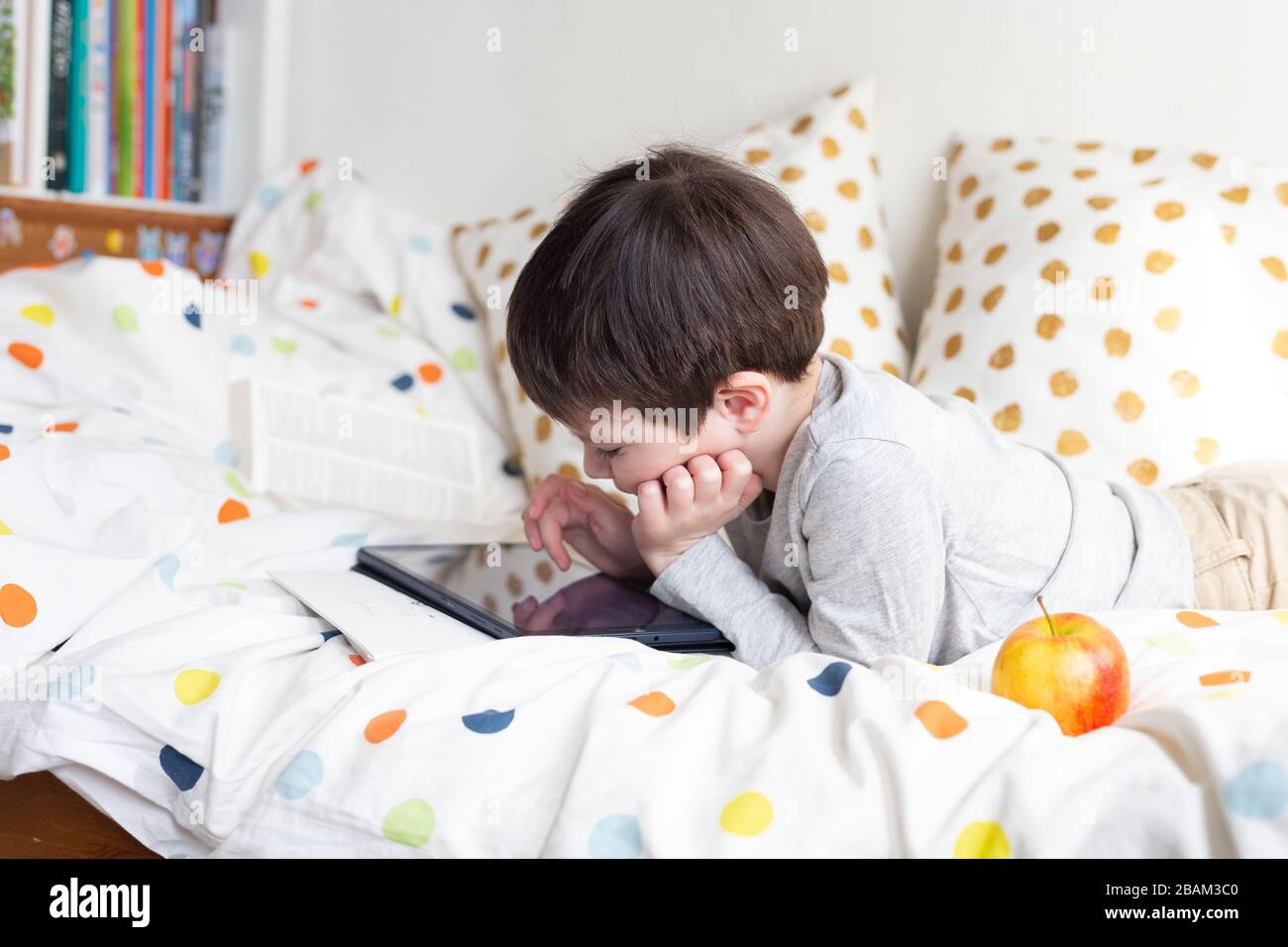 Scuola ragazzo a letto a casa con un tablet digitale in mano, facendo i compiti. Formazione online a distanza. Quarantena Foto Stock