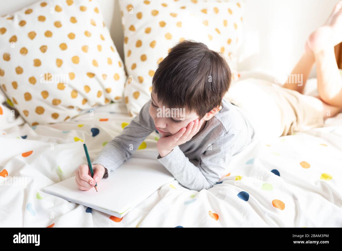 Ragazzo della scuola a letto e fare i compiti della scuola a casa, scrivendo in quaderno. Formazione online a distanza. Quarantena Foto Stock