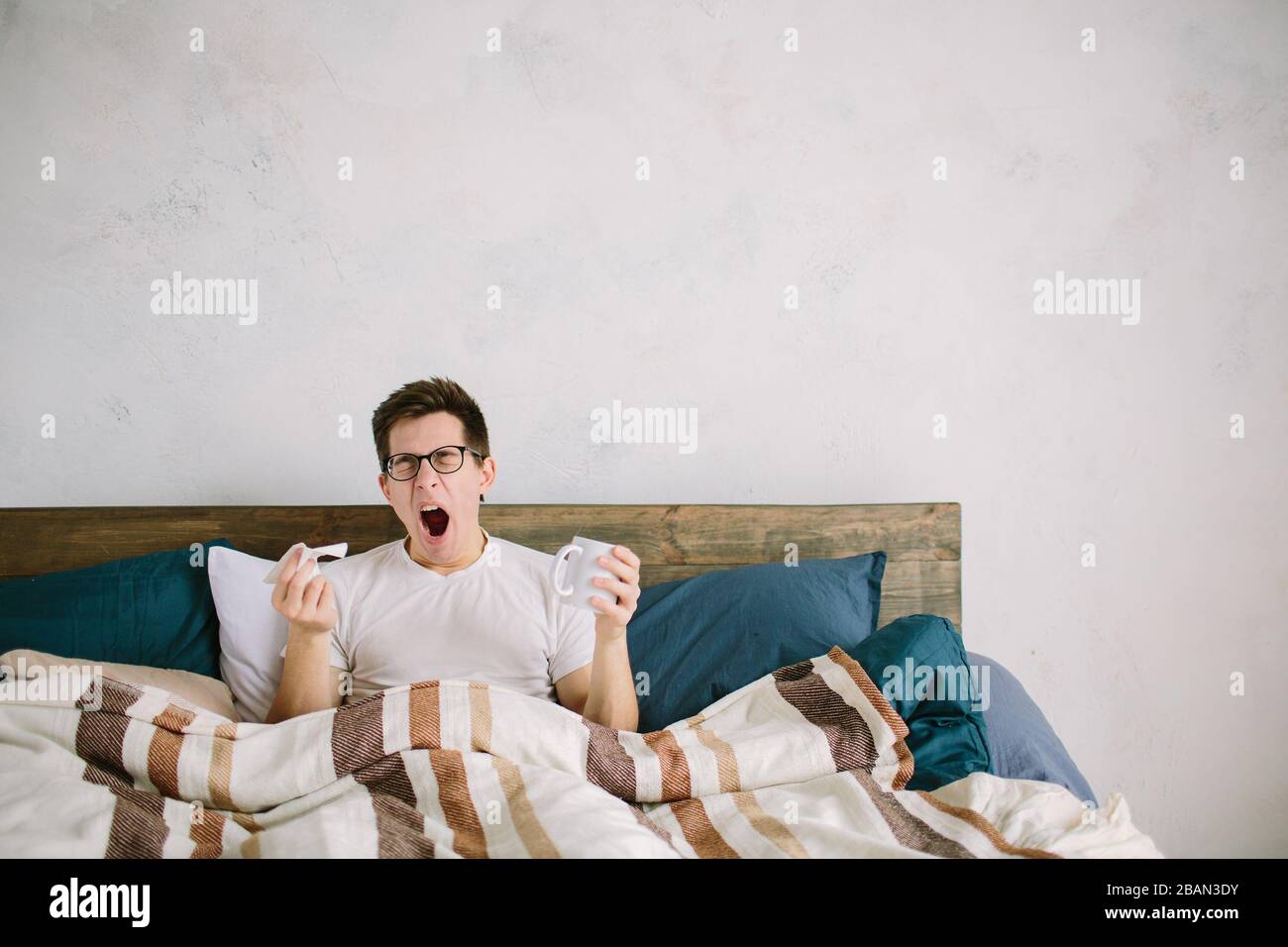 Mattina. L'uomo si trova a letto. Tiene in mano un tovagliolo e una tazza di tè o caffè. Il ragazzo non ottiene abbastanza sonno, sbadigia e si sente male. È caduto Foto Stock