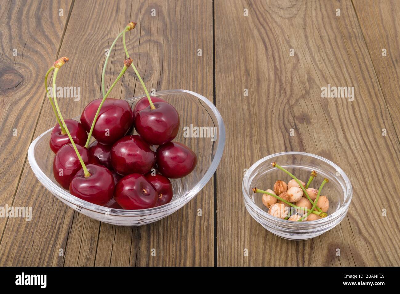 Mucchio di ciliegie rosse succose con steli verdi in vaso di vetro su sfondo di legno. Prunus avium. Primo piano di frutti dolci maturi e di ciliegie in piatto rotondo. Foto Stock