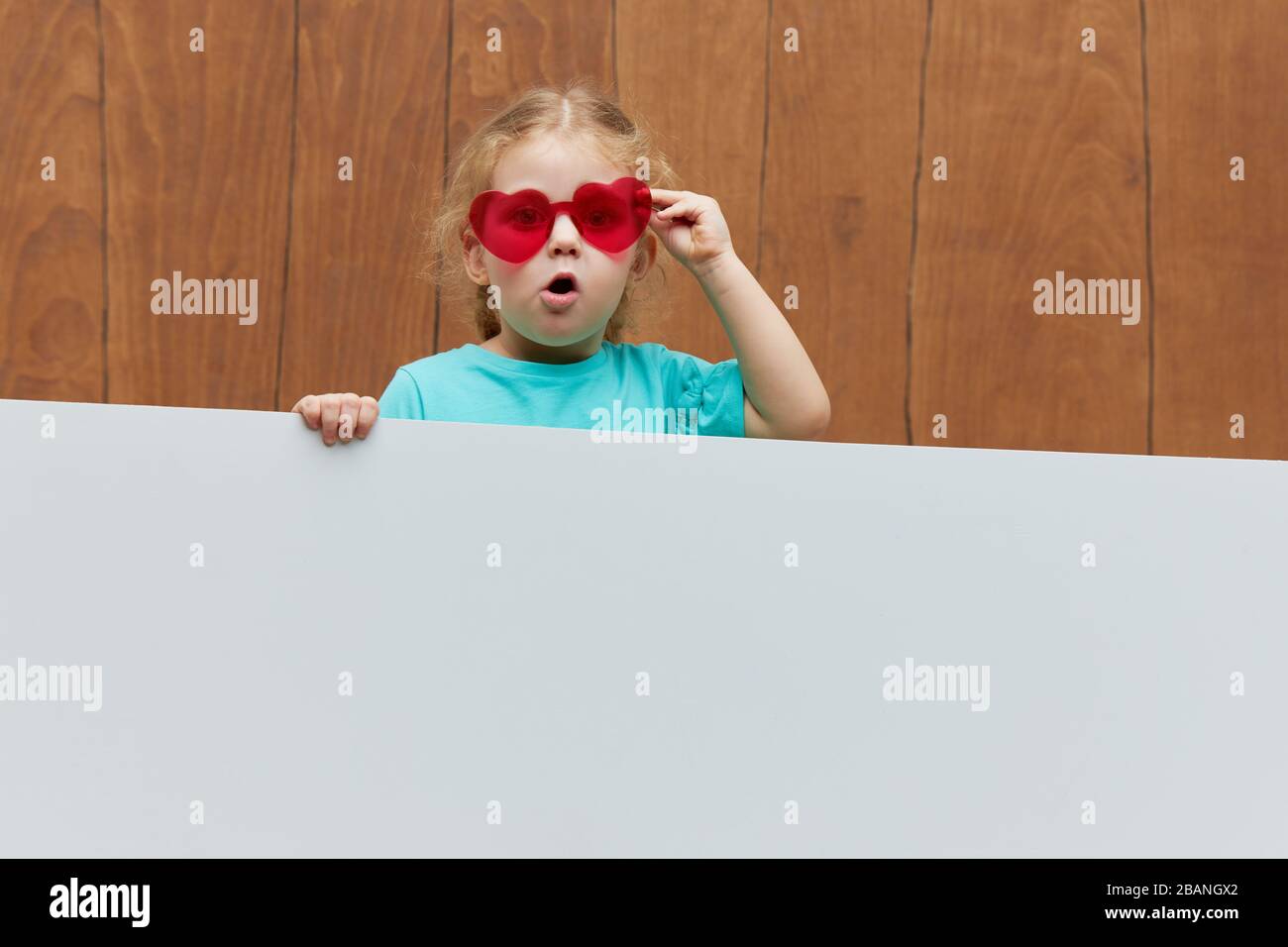 Piccoli occhiali a forma di cuore sorridenti ragazza bambino stando dietro un pannello bianco vuoto su sfondo di legno. Faccia divertente. Peeking fuori da dietro una ba Foto Stock