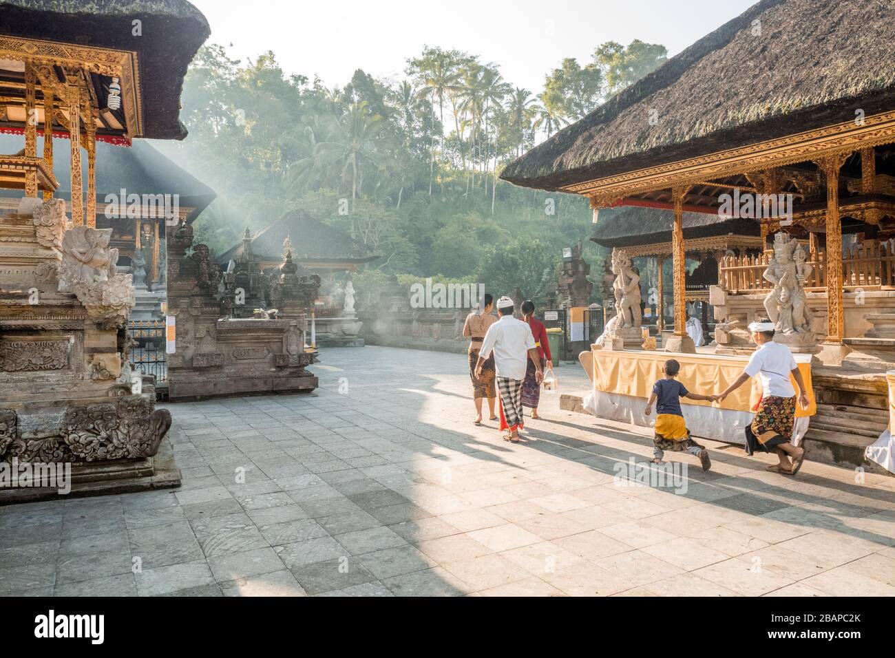 Famiglia Balinese in abito tradizionale che cammina attraverso il cortile del tempio in prima mattina luce misteriosa con fratelli che tengono le mani gettando lunghe ombre. Foto Stock