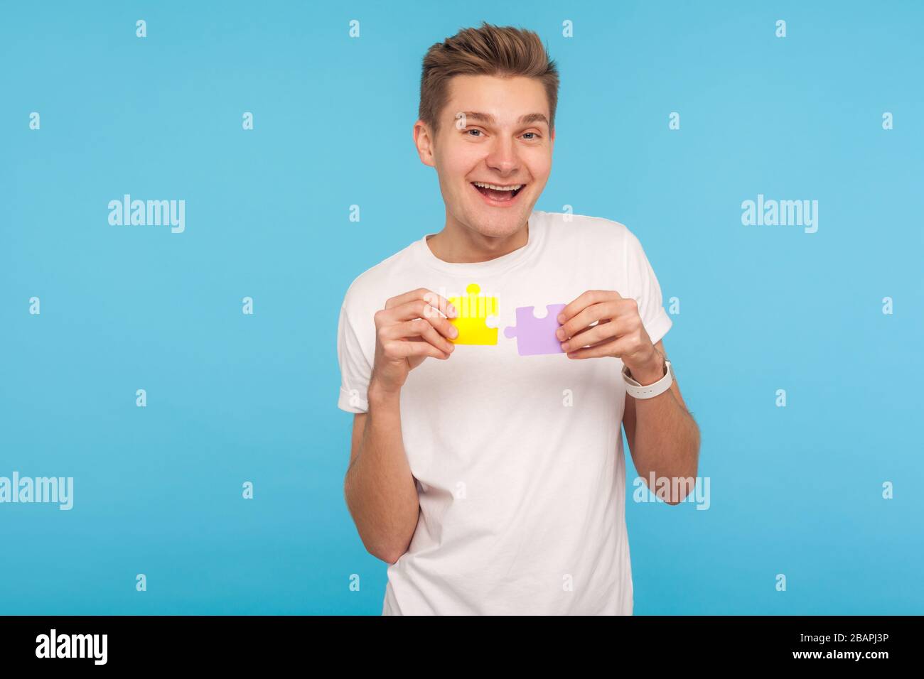 Uomo divertente positivo in t-shirt bianca con due parti di puzzle e sorridente gioiosamente, pronto a collegare i pezzi di puzzle, simbolo di Unione e associazione. In Foto Stock