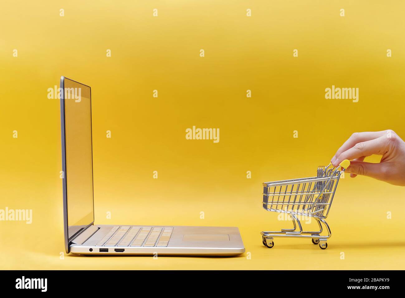 Tieni a mano il mini carrello per lo shopping vicino al laptop aperto su sfondo giallo. Foto Stock