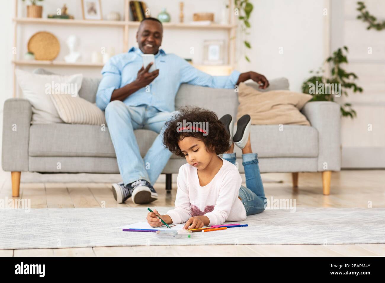 Resta a casa. Ragazza afroamericana che disegna sul pavimento mentre il suo nonno usando lo smartphone all'interno Foto Stock