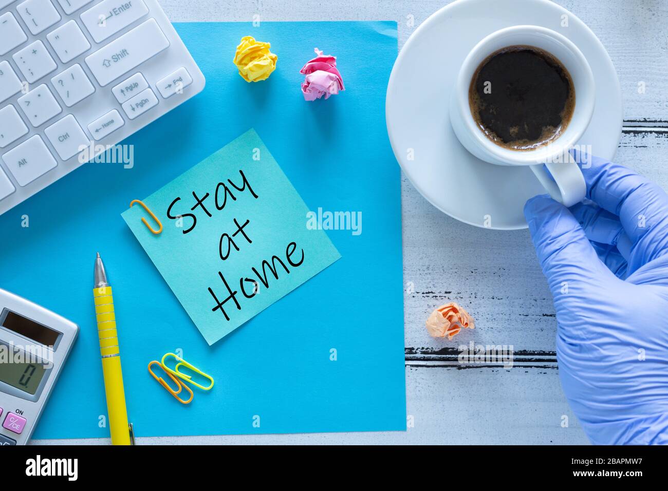 Carta con le parole 'Say at Home' mano in un guanto di gomma raggiungere per il caffè, spazio ufficio durante l'epidemia di Coronavirus Foto Stock