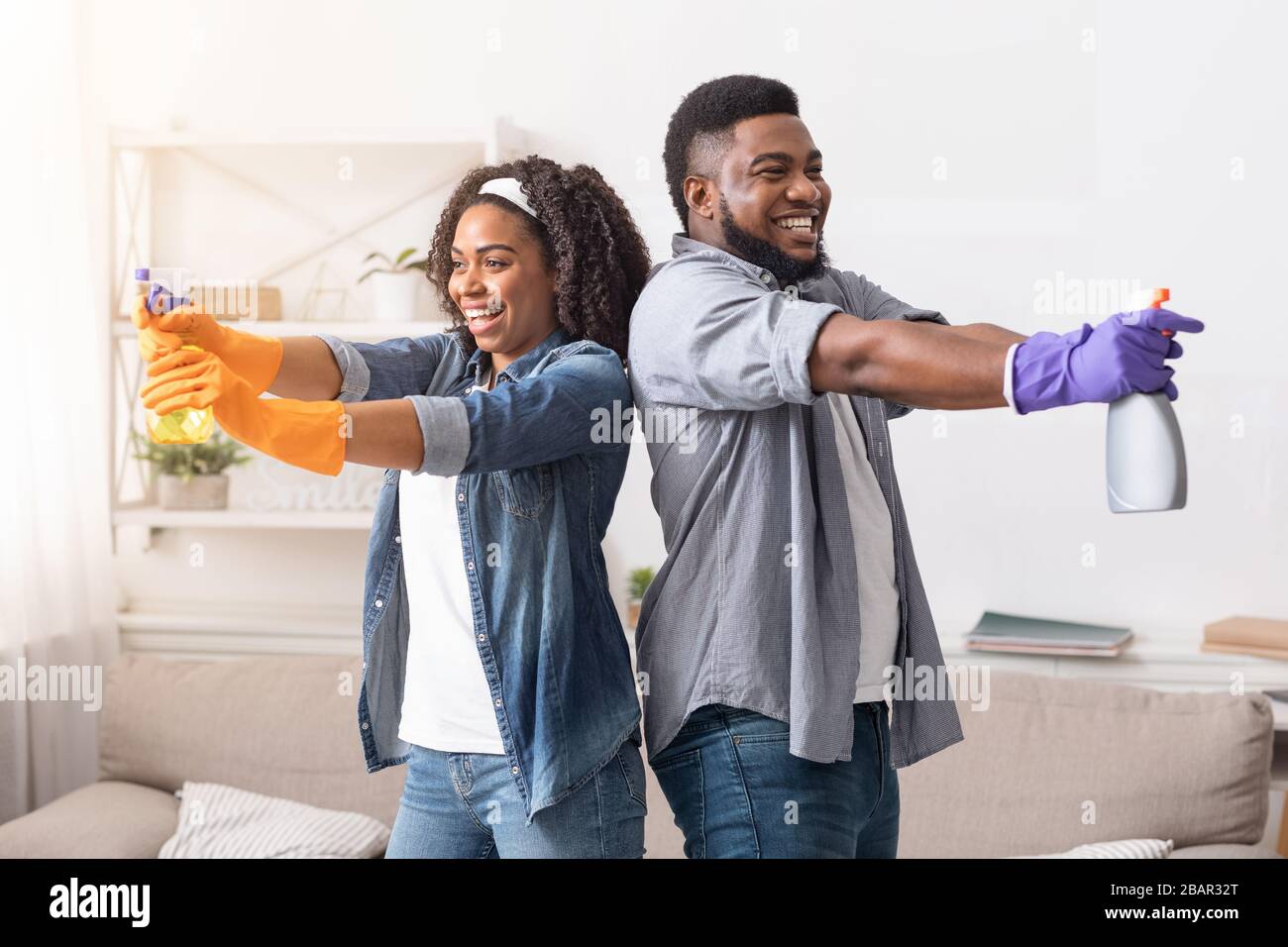 Coppia afro-americana che si divertono durante la pulizia dell'appartamento, puntando con detergenti spray e ridendo Foto Stock