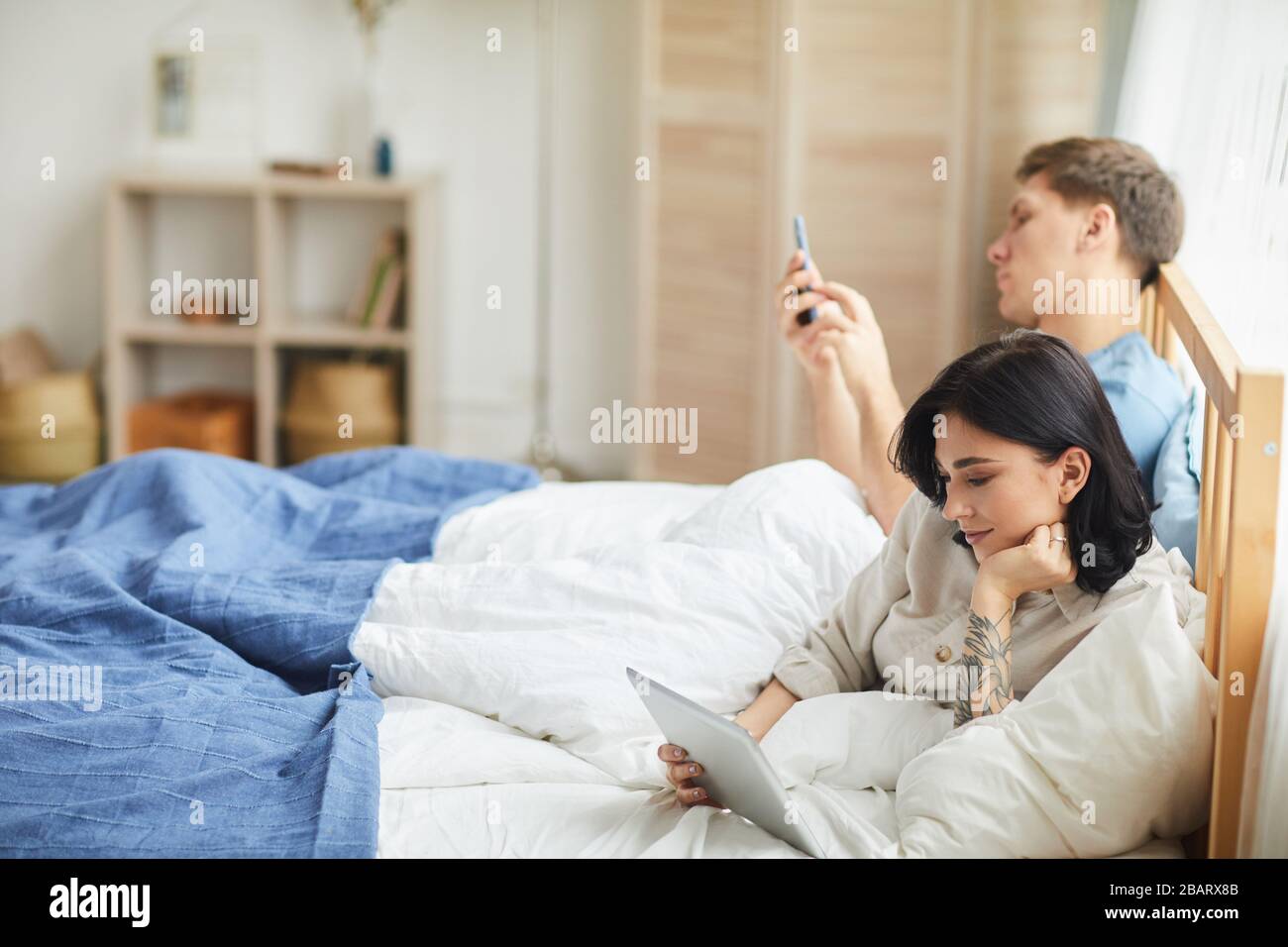 Ritratto di vista laterale di coppia moderna utilizzando dispositivi mobili e navigare in internet mentre sdraiato a letto al mattino, copia spazio Foto Stock
