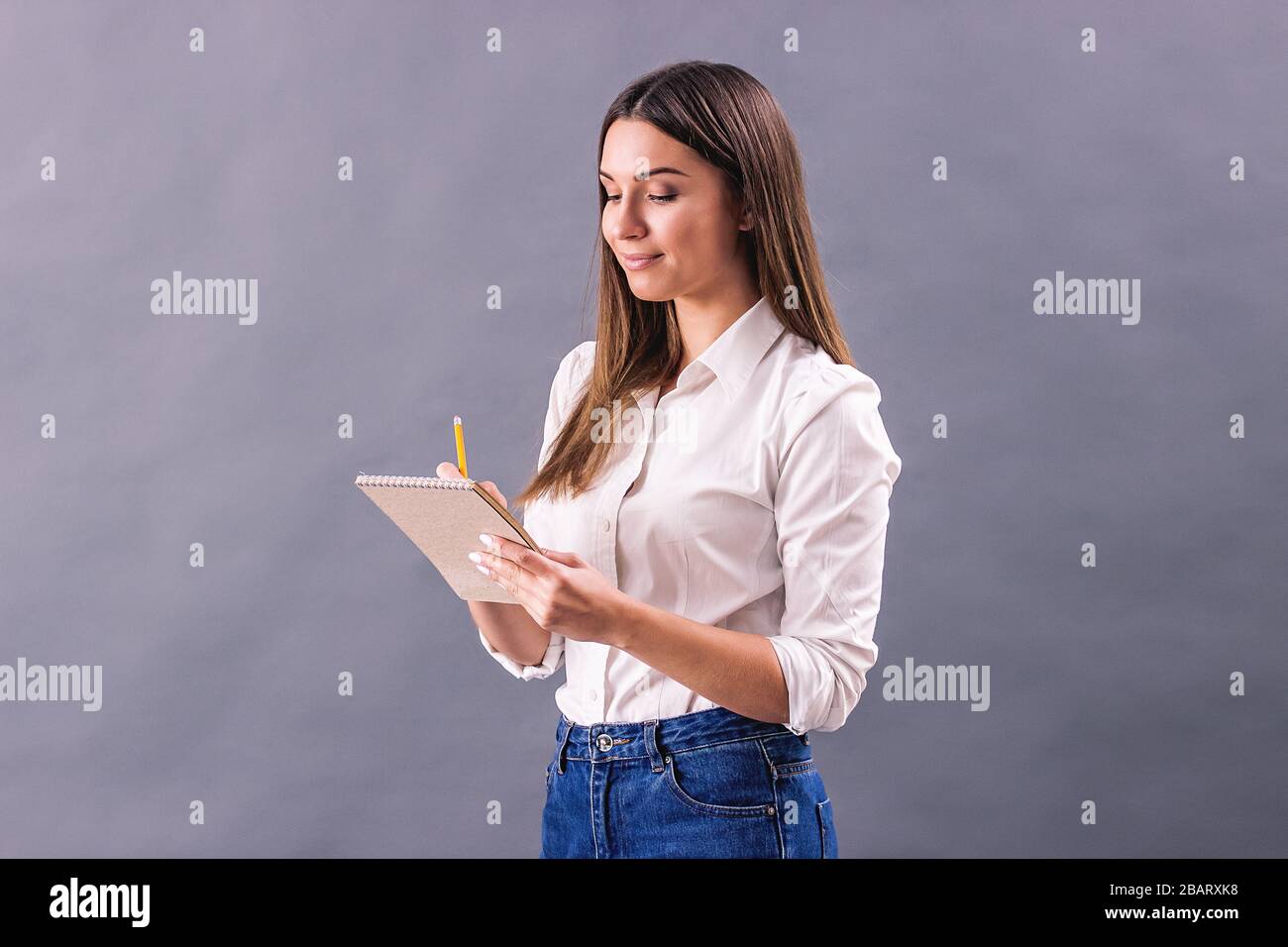 Giovane donna creativa che prende appunti nel suo notebook e sorridente mentre si sta isolati su sfondo grigio. Bella ragazza scrivere o disegnare schizzi i Foto Stock