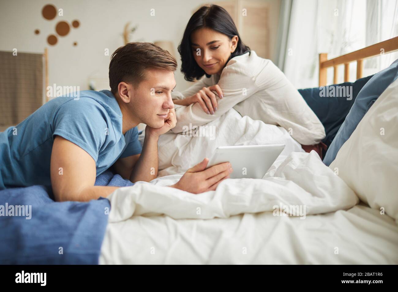 Ritratto di giovane coppia moderna usando il tablet digitale mentre si siede a letto a casa e ordinare cibo online o guardare film, copia spazio Foto Stock