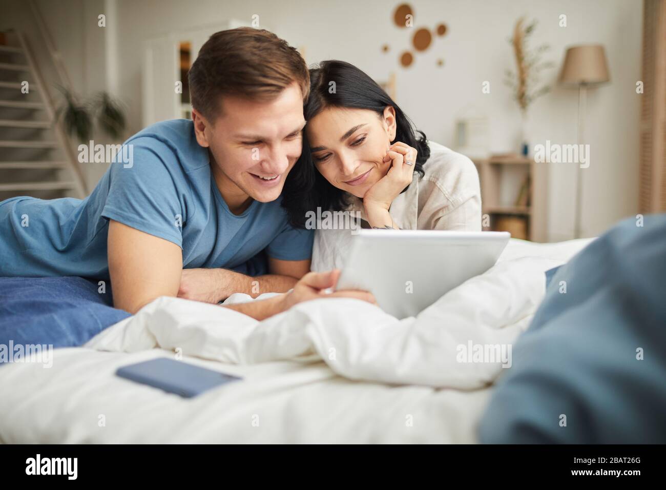 Ritratto di giovane coppia felice usando il tablet digitale mentre sdraiato a letto a casa e ordinare cibo online o guardare film, copia spazio Foto Stock