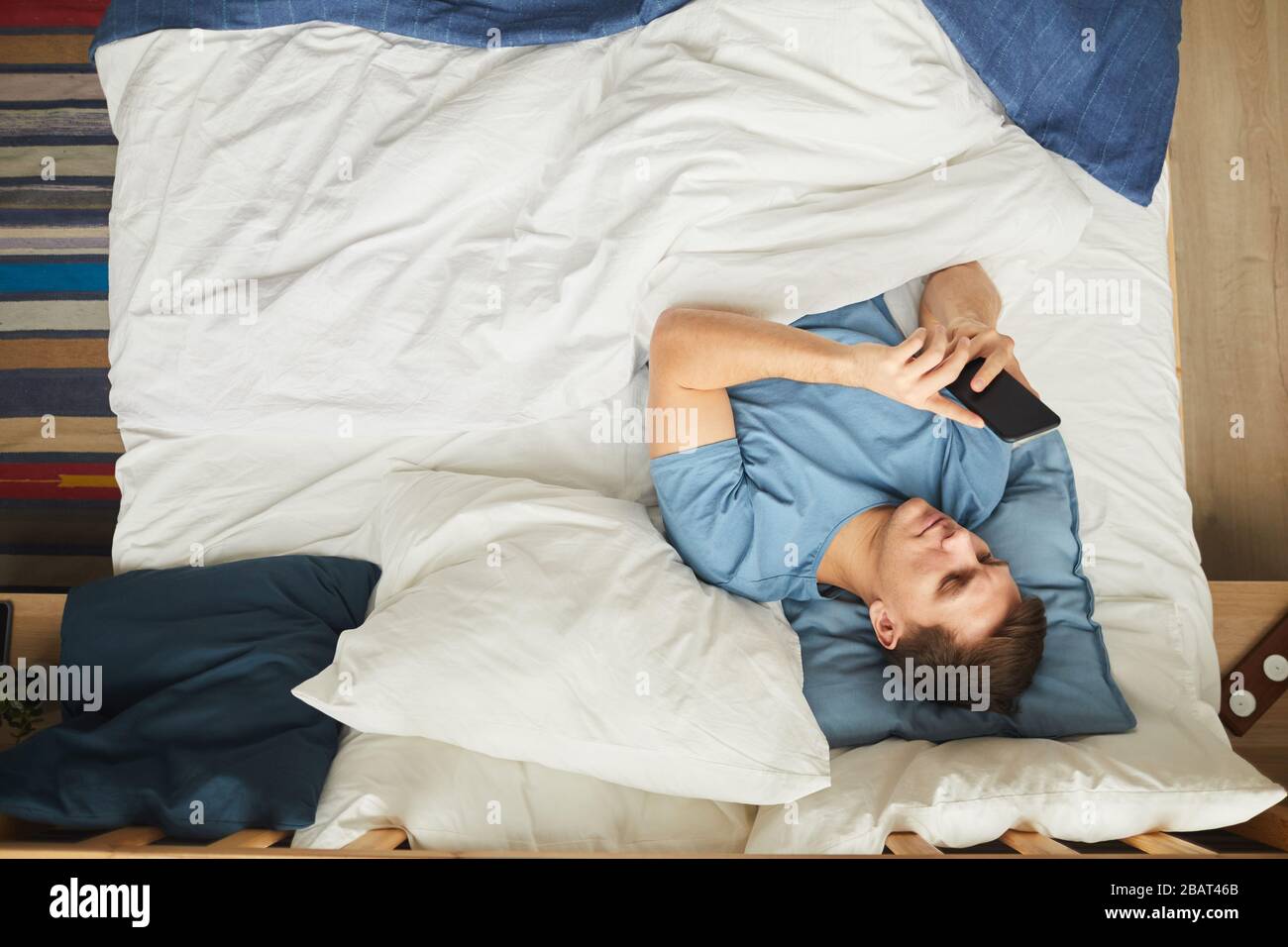 Sopra la vista ritratto di un giovane moderno utilizzando smartphone mentre sdraiato sul letto in blu e bianco interno, copia spazio Foto Stock
