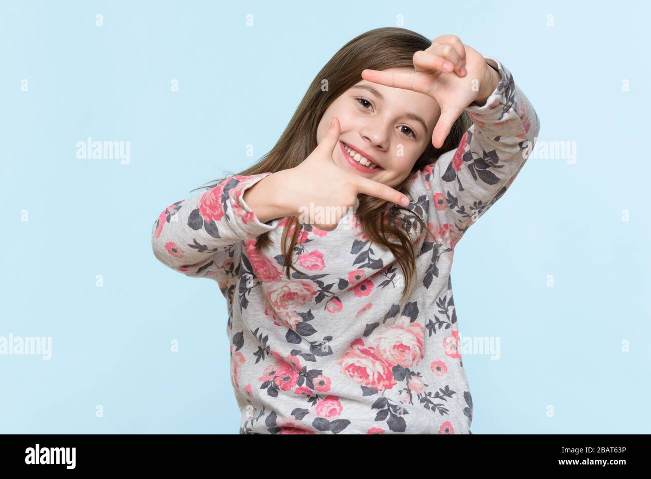 Adorabile ragazza di età elementare sorridente guardando la fotocamera e facendo foto cornice gesto mano. Monolocale su sfondo blu pastello. Foto Stock