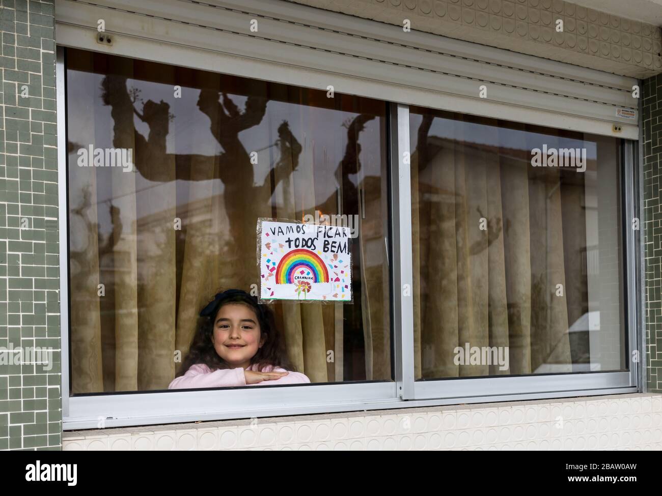 Piccola ragazza alla finestra della sua casa (Portogallo) con un dipinto arcobaleno che dice 'We All will be alright' sotto la campagna di soggiorno di COVID-19 Foto Stock