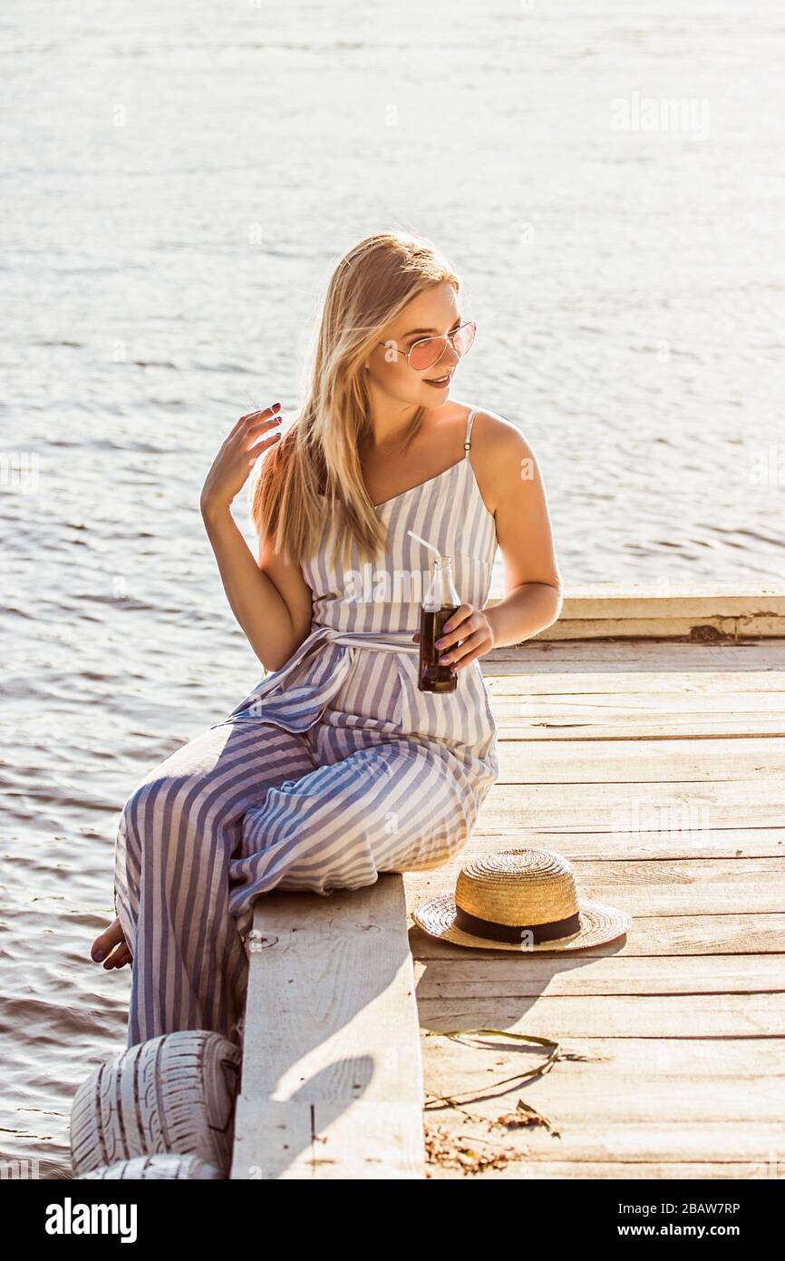 Dolce ragazza attraente in elegante abbigliamento estivo si siede sul molo  di legno sulla spiaggia e bevande soda da bottiglia. Bella giovane bionda  donna in spogliata Foto stock - Alamy