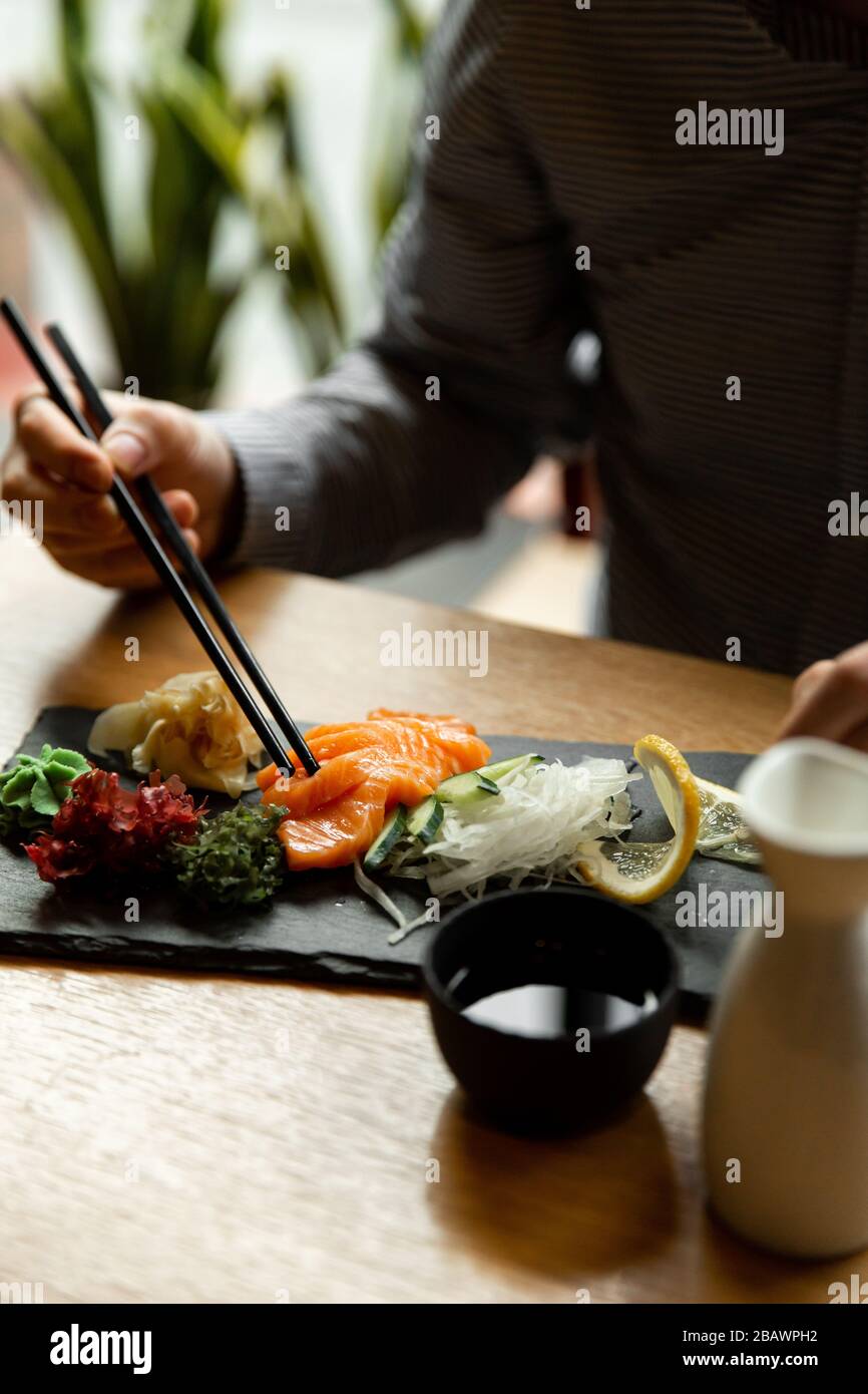 Uomo che tiene un bastone con sashimi di salmone sul tavolo del ristorante. L'uomo asiatico mangia sashimi in un ristorante asiatico. Foto Stock