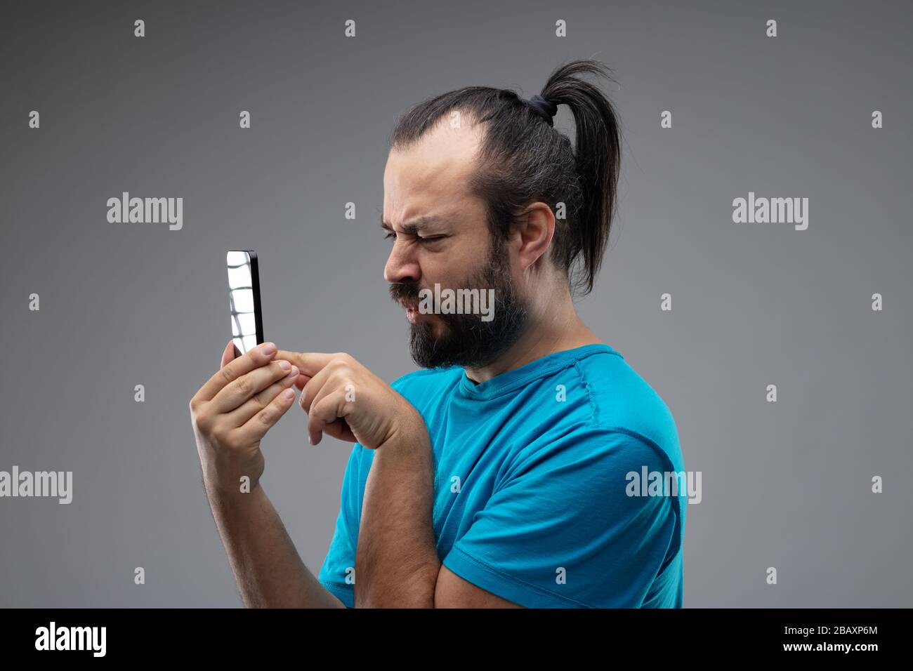 Uomo con bearded in t-shirt blu, con capelli neri pettinati in coda di cavallo, guardando lo smartphone, tenendo in mano vicino al suo viso, come se si avendo cattiva vista. Laterale Foto Stock