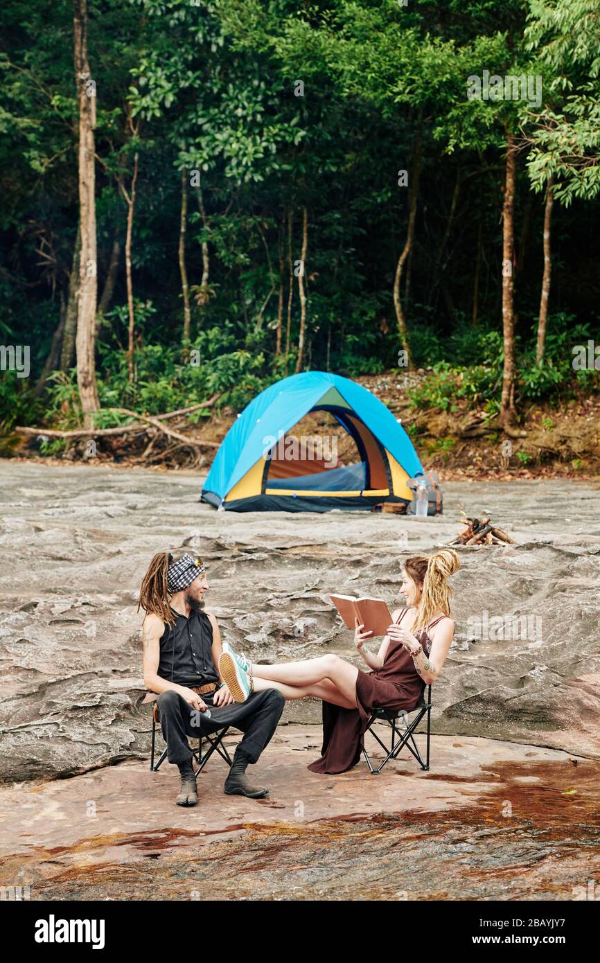 Donna che mette le gambe su giri di marito e di lettura lui un libro quando sono campeggio in foresta Foto Stock