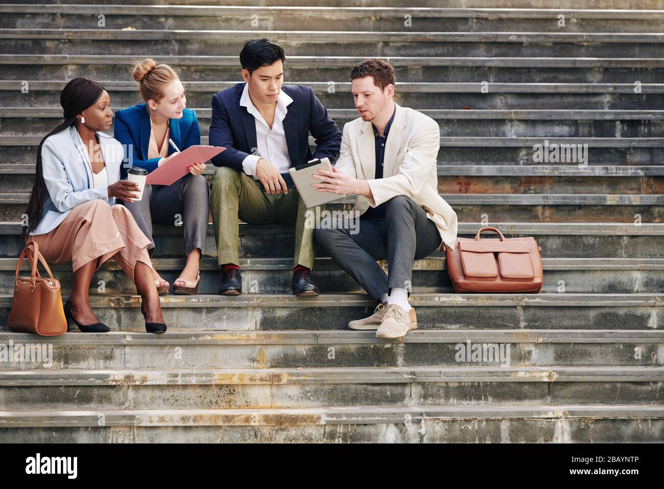 Gruppo di persone aziendali seri che discutono nuove tendenze e tendenze quando si siedono su gradini e guardano video su tablet digitali Foto Stock