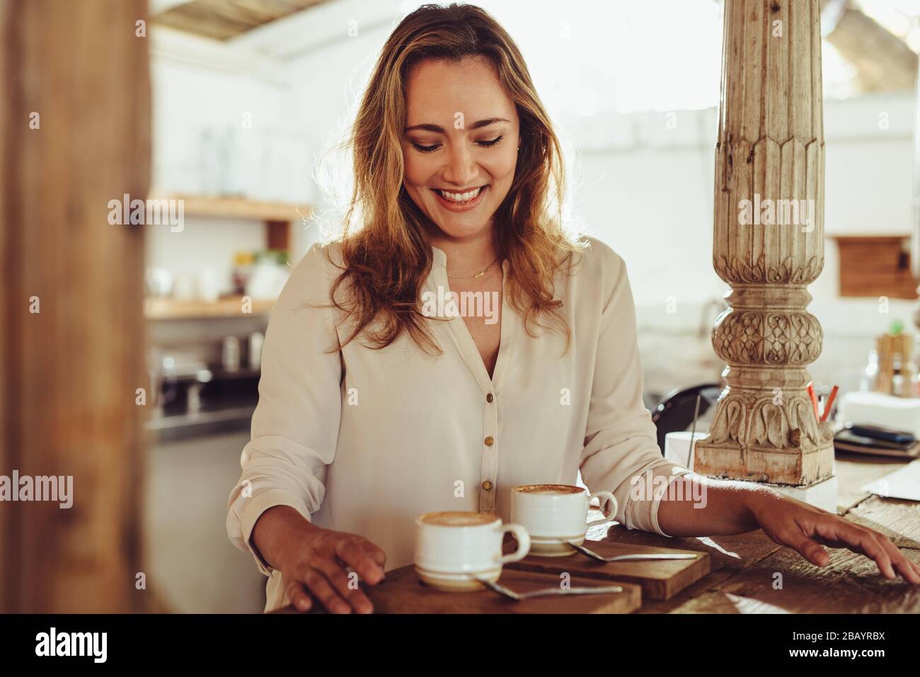 Donna sorridente che si trova dietro il bancone della caffetteria di una caffetteria. Barista femminile sorridente che serve due tazze di caffè. Foto Stock