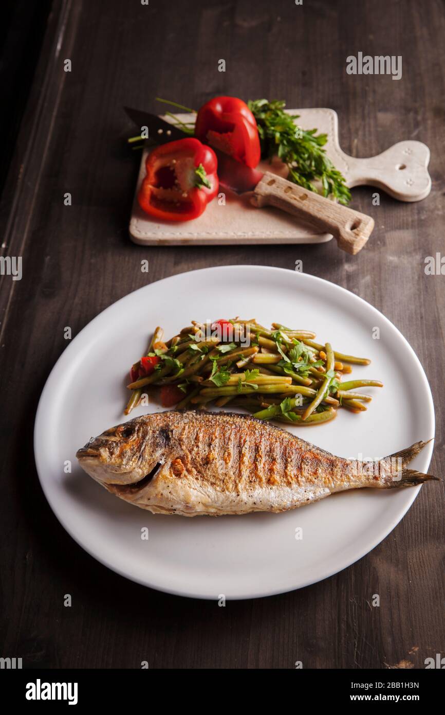 pesce dorado grigliato mediterraneo con fagioli verdi al vapore su piatto bianco su tavolo di legno scuro Foto Stock
