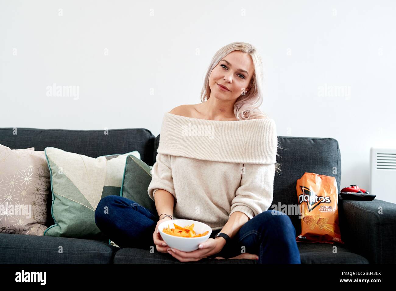 Una singola femmina caucasica bionda seduta su un divano con una ciotola bianca di formaggio piccante Doritos Foto Stock