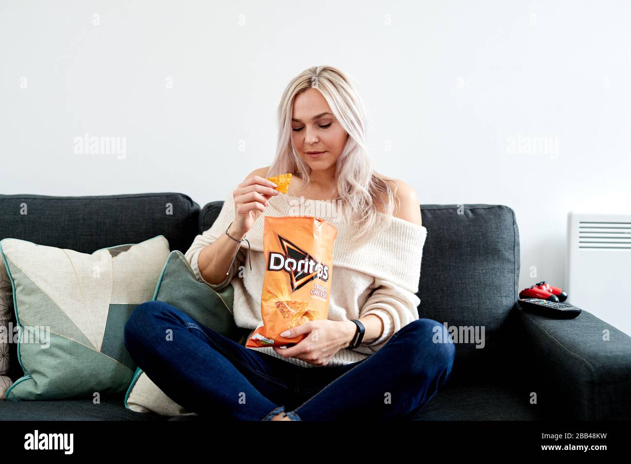 Una singola femmina caucasica bionda seduta su un divano guarda in una borsa di formaggio piccante Doritos Foto Stock