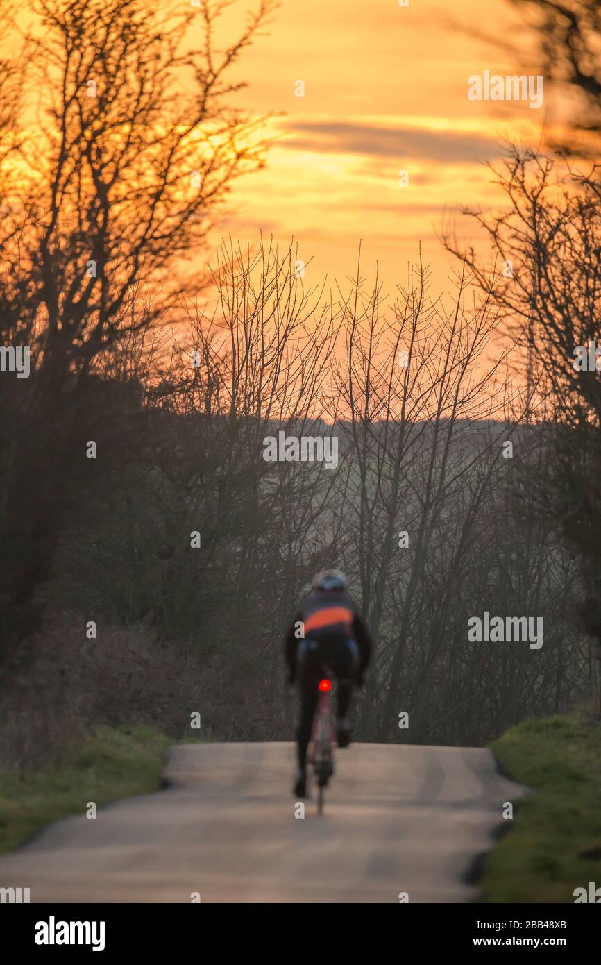 Tramonto cielo sera in campagna rurale; vista posteriore di un ciclista isolato a cavallo della sua bicicletta lungo la strada vuota. Uomo in bicicletta con casco da dietro. Foto Stock