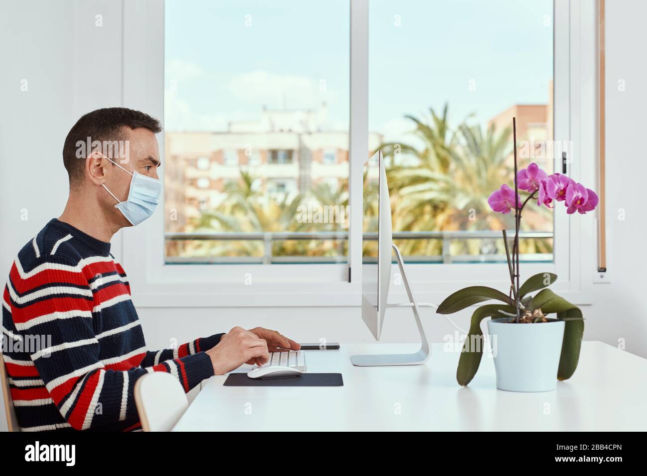 primo piano di un giovane caucasico all'interno, con una maschera chirurgica, con un computer seduto su un tavolo bianco Foto Stock