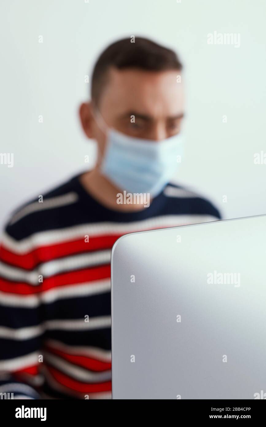 un giovane caucasico all'interno, indossando una maschera chirurgica, usando un computer, con il fuoco nel computer in primo piano Foto Stock