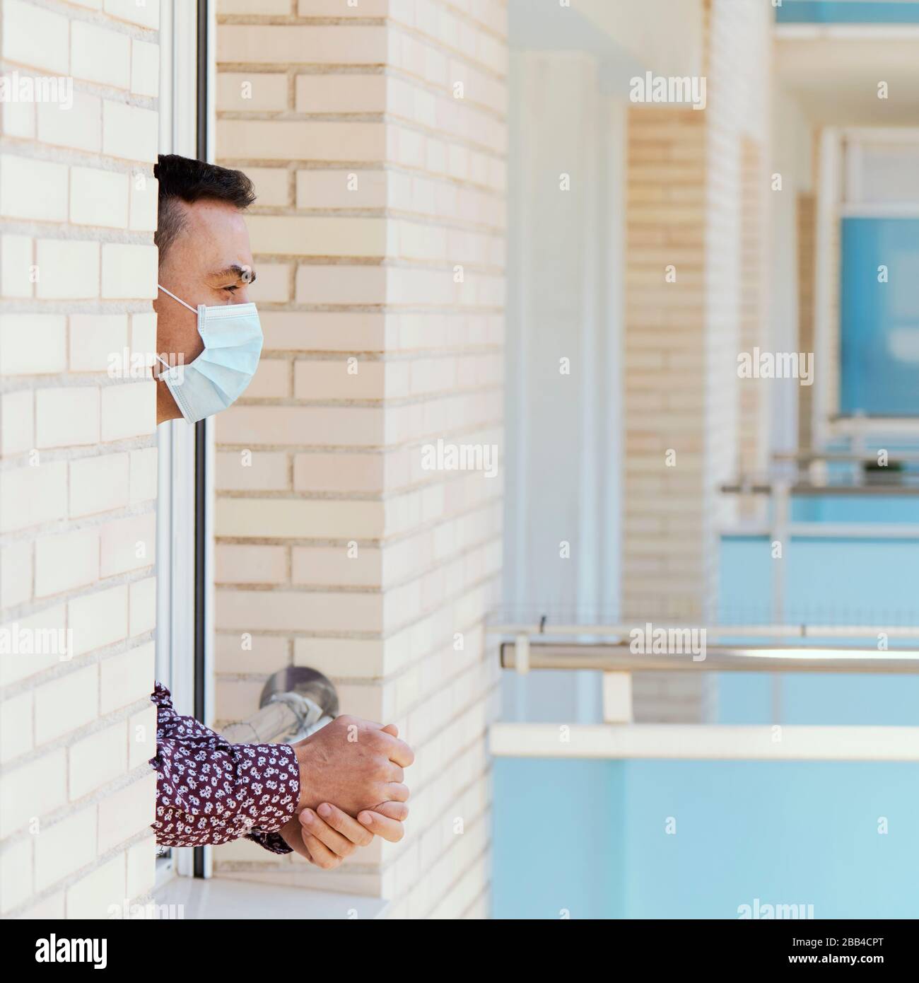 vestirsi di un giovane caucasico indossando una maschera chirurgica appoggiata nella finestra della sua casa o di una camera d'albergo Foto Stock