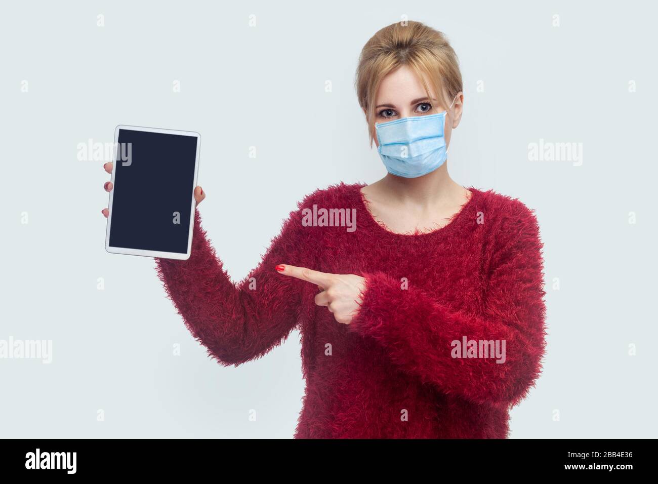 Ritratto di bella giovane donna con maschera medica chirurgica in blusa rossa in piedi e tenendo compressa schermo vuoto e punta il dito verso il dispositivo e. Foto Stock