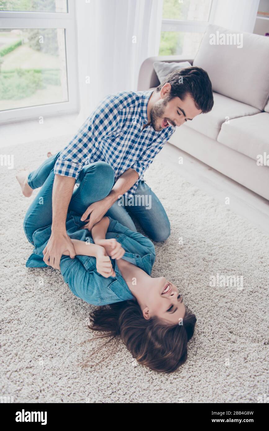 La giovane coppia eccitata sta ingannando intorno e gioca all'interno a casa, ad un tappeto beige accogliente, indossando abiti casual, ridendo Foto Stock