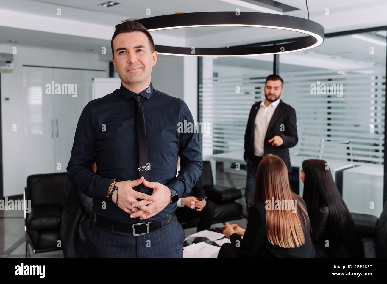 Fiducioso giovane tenendo le braccia incrociate mentre permanente al tavolo parlando con i colleghi su uno sfondo Foto Stock