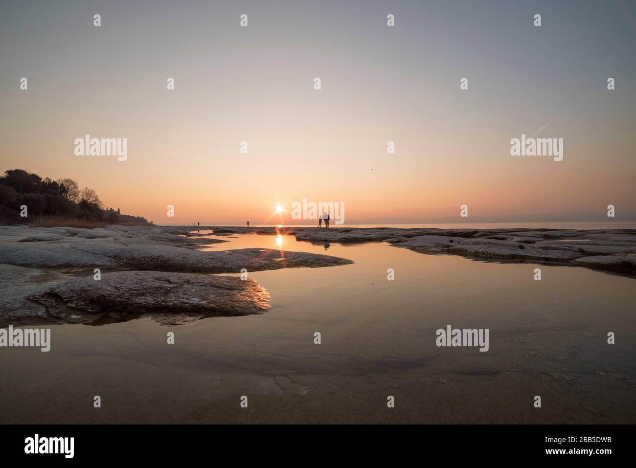 spiaggia di sirmione di notte e tramonto sul lago di Garda con rocce piatte sulla spiaggia e acqua e sole in l'orizzonte Foto Stock