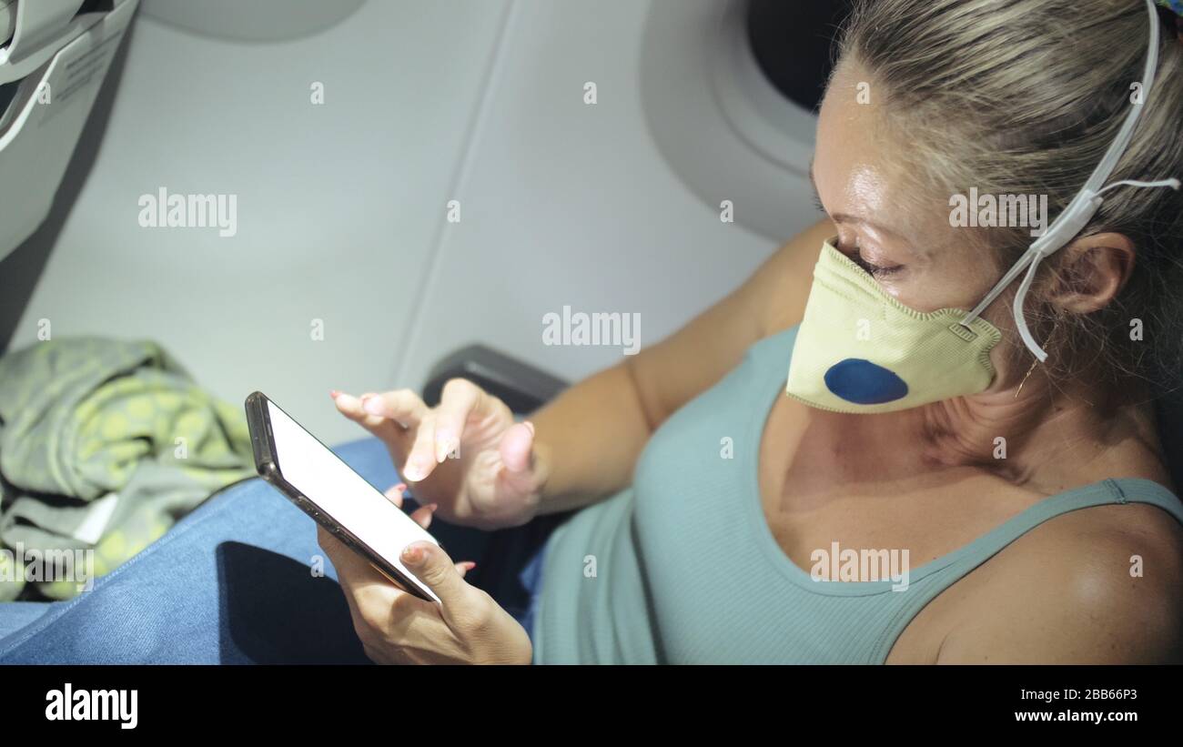Donna viaggio turistico caucasico in aereo aereo con indossare maschera medica protettiva. Leggi le notizie sugli shock relativi a coronovirus su smartphone mobile. Foto Stock