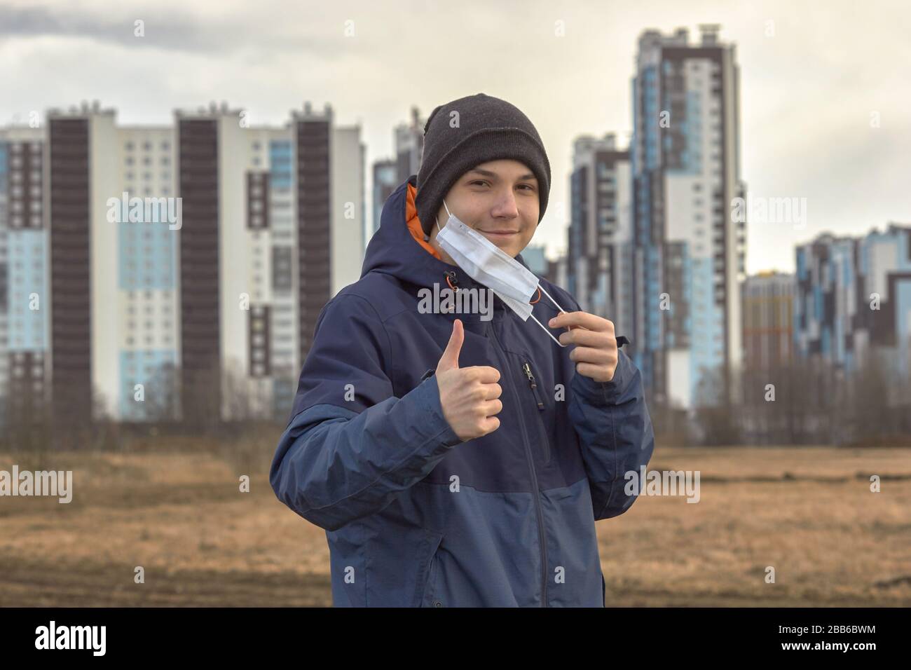 un adolescente in abiti scuri demi-stagione toglie una maschera medica contro lo sfondo di alti blocchi Foto Stock