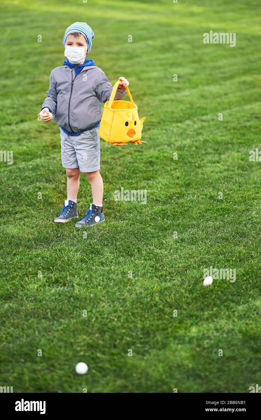 Ragazzino che indossa maschera protettiva alla caccia dell'uovo di Pasqua nel giardino primaverile. Foto Stock