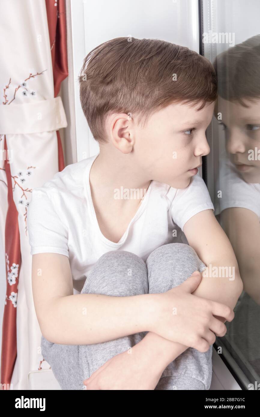 Un ragazzo solitario di sette anni si siede su un davanzale, guarda fuori la finestra ed è triste. Autoisolamento, concetto di quarantena del caronavirus Foto Stock